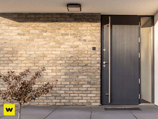 Moderne Haustür aus dunklem Material in Ziegelsteinfassade mit kleinem Baum davor.