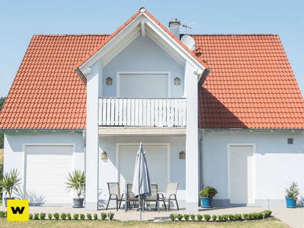 Modernes Einfamilienhaus mit rotem Dach, Terrasse und geschlossenen Rollläden bei Sonnenschein.